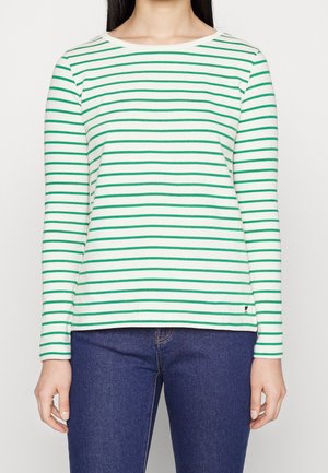 Vrouw met een wit langemouwenshirt met groene horizontale strepen en blauwe jeans, staand tegen een effen lichte achtergrond.