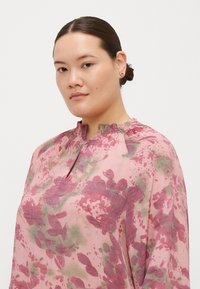 Blusa a fantasia floreale in morbido tessuto rosa con dettagli verdi, caratterizzata da un colletto arricciato e un'apertura a chiave. Design leggero e texturizzato.