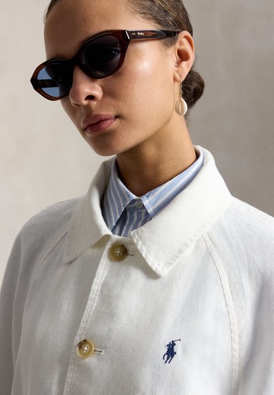 Femeie purtând ochelari de soare Polo în culoarea scoicii de broască țestoasă închisă, cercei picătură din scoici, jachetă albă cu guler și cămașă în dungi albastre dedesubt.