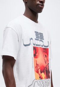 Άνδρας που φοράει λευκό μπλουζάκι με αφηρημένο κόκκινο σχέδιο και γκρι στιλιζαρισμένο κείμενο μπροστά, με απλό γκρι φόντο.