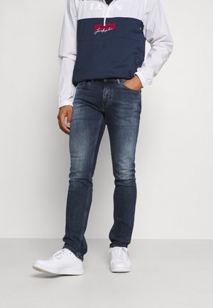 Man wearing dark blue jeans, white sneakers, and a white and navy blue Hollister jacket with red logo, standing with one hand in pocket.