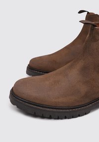 Botas de tobillo de ante marrón con puntera redondeada, tacón bajo y suela de goma texturizada. Presenta tiradores para facilitar el uso y costuras en contraste.