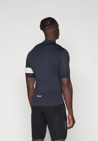 Maillot de cyclisme marine en tissu extensible, avec des manches courtes, une bande d'accent blanche et une poche arrière. Associé à un short de cyclisme noir.