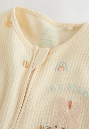 Light yellow ribbed fabric onesie with a front zipper, featuring embroidered bunny, rainbow, and text “MY FIRST.” Soft texture with playful details.