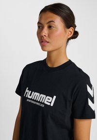 Sort Hummel t-shirt lavet af bomuld, med et fremtrædende hvidt logo og en klassisk rund halsudskæring, samt hvide chevron-detaljer på ærmerne.