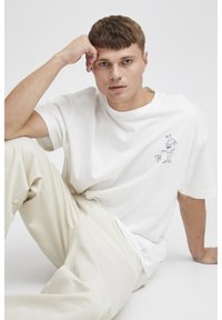 Hvid bomulds-T-shirt med rund halsudskæring, der har en minimalistisk blå grafisk illustration på brystet. Sammen med beige bukser.