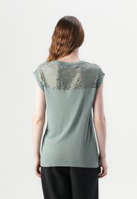 Krátký rukáv top v tlumené zelené barvě s texturovaným krajkovým výstřihem. Materiál je hladký a lehký, má kulatý výstřih a uvolněný střih.