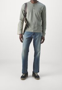 Långärmad grön henley-tröja med tre knappar, kombinerad med blå raka jeans och svarta sneakers med bruna sulor, bärande en beige axelväska.