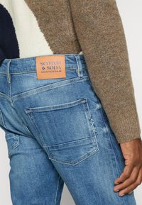 Středně modré džínové kalhoty s hnědou koženou cedulkou "SCOTCH & SODA AMSTERDAM", zakládající se na detailech prošívání zadní kapsy a klasickém střihu.