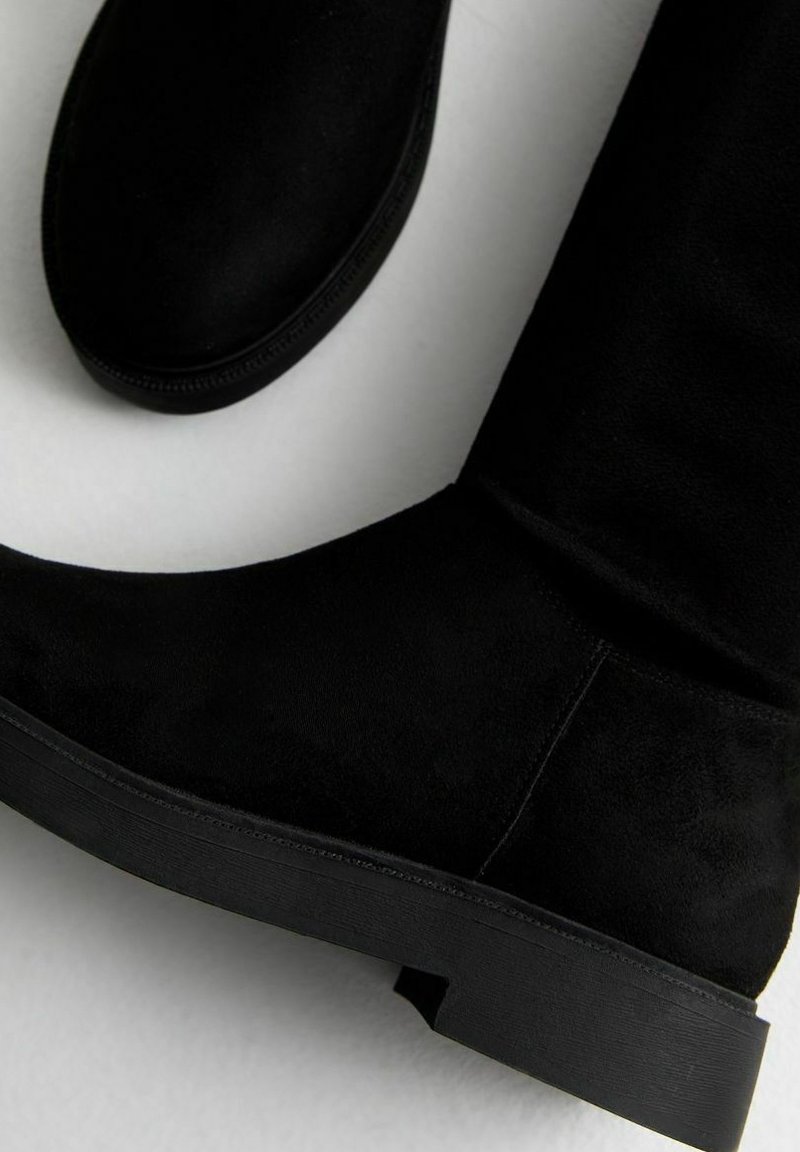 Schwarze Wildleder-Stiefel bis zum Knie mit flacher Gummisohle und nahtlosem Design. Der Schaft ist strukturiert mit einer glatten Oberfläche und ohne sichtbare Beschläge.