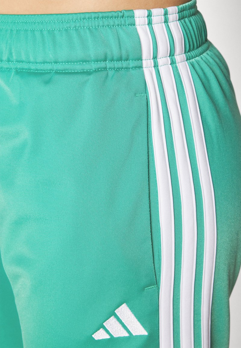 Pantalones deportivos en color turquesa con franjas laterales blancas, cinturilla elástica y un logotipo triangular bordado en la parte baja de la pierna. Textura suave.