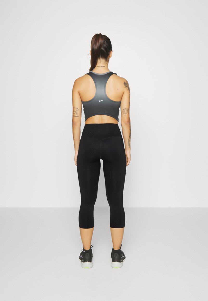 Soutien-gorge de sport gris avec un dos découpé et un logo, associé à un legging noir long jusqu'à la cheville. Le modèle porte des baskets noires avec des accents verts.