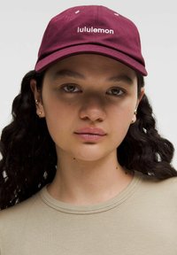 Cappellino bordeaux con logo "lululemon" bianco, visiera curva e sei fori di ventilazione. Tessuto liscio con design semplice. Il modello indossa una maglietta beige.