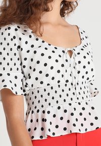 Mujer vestida con una blusa blanca con lunares negros, cintura y mangas fruncidas, y detalle de lazada en el escote, combinada con pantalones rojos.