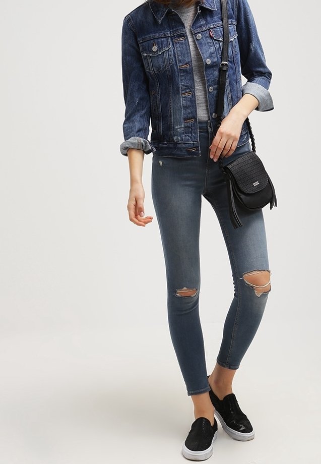 Femme portant une veste en jean, un jean skinny déchiré, des chaussures noires à enfiler et un sac bandoulière noir, debout devant un fond uni.