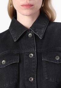 Giubbotto in denim nero con colletto a punta, bottoni a pressione argentati e due tasche sul petto. Il tessuto presenta una trama sottile e dettagli di cucitura visibili.