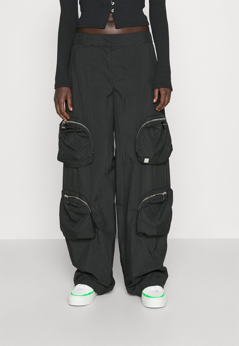 Zwarte cargo pants van lichtgewicht stof, voorzien van meerdere met rits afgesloten zakken en een ontspannen pasvorm. Gepaard met witte schoenen met groene accenten.