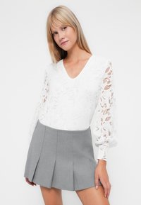 Blusa bianca in pizzo con scollatura a V e maniche a sbuffo, abbinata a una mini gonna a pieghe grigia aderente. Texture morbide e colori a contrasto.