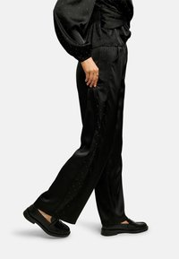 Sieviete, kas valkā melnas zīda bikses un piemērotu topu ar maziem baltiem punktiem, melnus loafer apavus, stāv sānis ar vienu kāju priekšā.