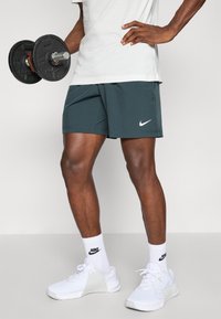 Pantalones cortos deportivos grises con un logo de Nike, combinados con zapatillas y calcetines atléticos blancos. El modelo sostiene una mancuerna negra.