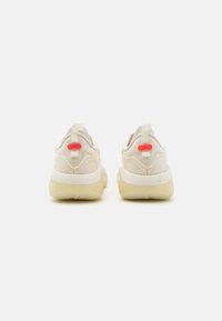 Beige sneakers med tekstureret mesh-overdel, gummisål og en trækflap. Har lyse røde detaljer på hælen for bedre synlighed.