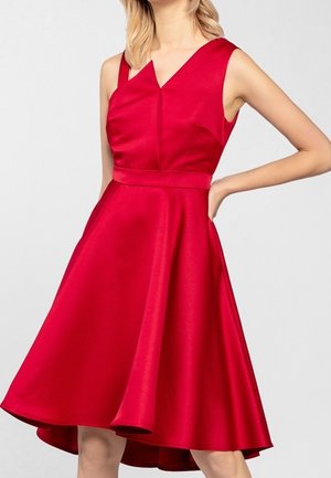 Femme portant une robe rouge vif sans manches, longueur genou, avec une taille cintrée et une jupe évasée sur un fond clair uni.