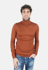 Uomo con capelli ricci e scuri che indossa un maglione marrone a collo alto e jeans blu, in piedi davanti a uno sfondo bianco semplice.