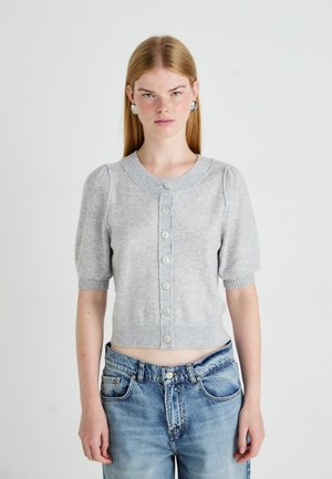 Young woman with long blonde hair wears a light gray buttoned short-sleeve sweater and blue jeans, standing against a plain background.