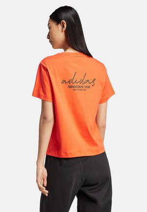 Oranges Baumwoll-T-Shirt mit kurzen Ärmeln, das auf der Rückseite schwarzen Text enthält, der "adidas FOUNDATION OF SPORT" in einer Schreibschrift darstellt.