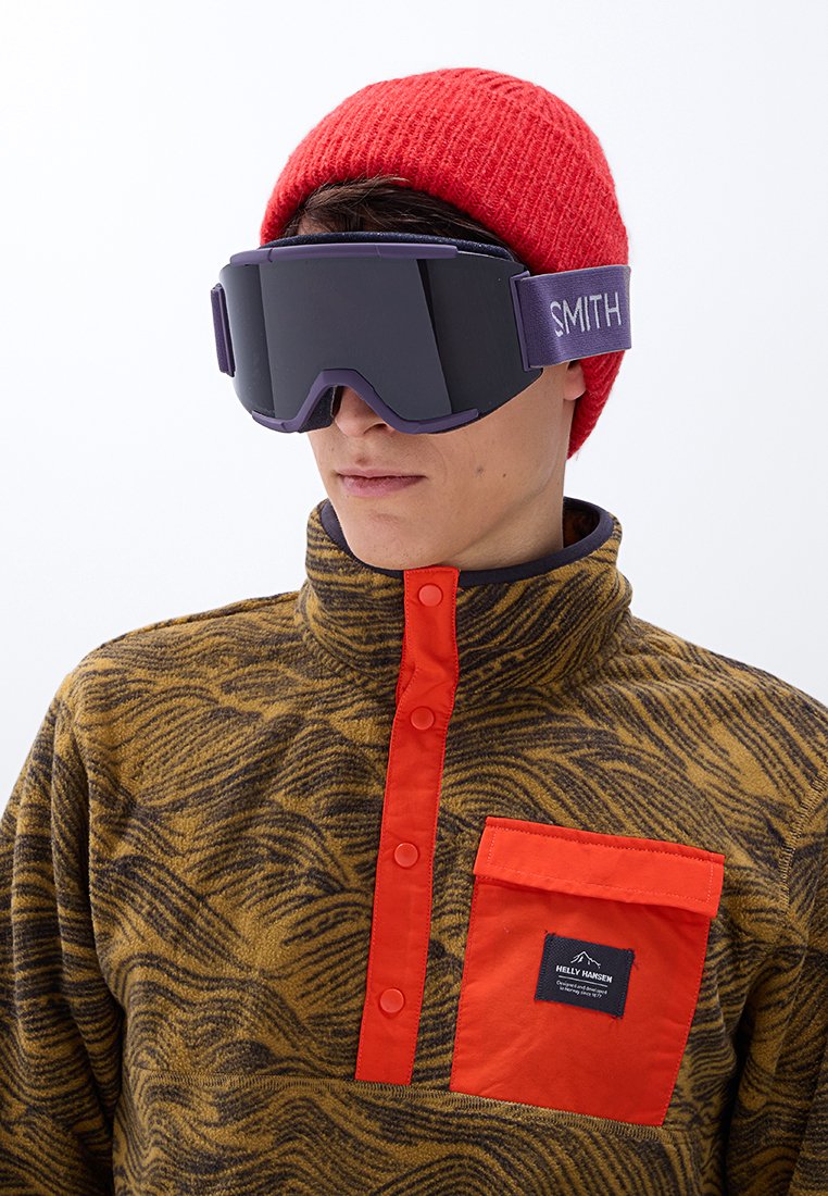 Ski-Brille mit großen schwarzen Gläsern, lila Riemen und Logo; Fleece-Pullover in Braun- und Gelb-Muster mit rotem Knopfverschluss und Tasche.