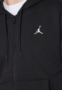 Felpa nera con zip, indossata sopra una maglietta bianca, con un piccolo logo Jumpman bianco ricamato sul lato sinistro del petto.
