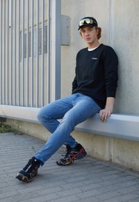 Fekete pulóver, kék farmer és fekete-narancs sportcipők, texturált talppal. Egy szürke padon ül, világos betonfal előtt.