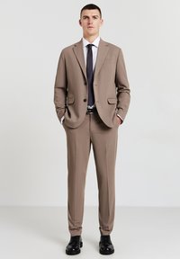 Completo sartorial beige con giacca monopetto, pantaloni abbinati, camicia bianca e cravatta nera. Scarpe stringate nere completano il look.