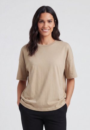 Kvinde med mørkt bølget hår iført en beige kortærmet skjorte og sorte bukser, smilende med hænderne i lommerne mod en hvid baggrund.