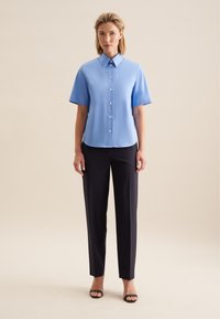 Jasnoniebieska koszula z krótkim rękawem, z kołnierzykiem, zapinana z przodu, w połączeniu z ciemnymi, dopasowanymi spodniami i czarnymi sandałami na obcasie na neutralnym tle.