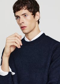 Dunkelblaues Strickpullover mit rundem Halsausschnitt, mit gerippten Bündchen und Saum, über einem weißen Hemd mit Kragen getragen. Schlichtes, strukturiertes Design.