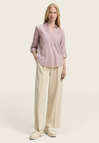 Woman standing with hands in pockets wearing a light pink button-up shirt, beige wide-leg trousers, and cream loafers against a plain background.