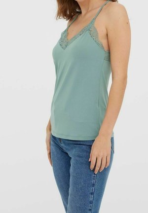 Jasnozielony top na ramiączkach z koronkowym wykończeniem przy dekolcie i regulowanymi ramiączkami, połączony z niebieskimi dżinsami. Gładka faktura materiału.
