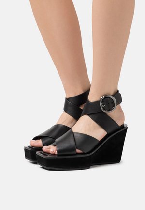 Mustat nahkaiset kiilakorkosandaalit leveillä ristiin menevillä remmeillä ja suurilla, pyöreillä soljilla, joita on käytetty paljain jaloin yksinkertaista vaaleaa taustaa vasten.