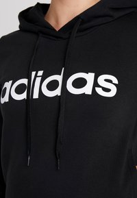 Felpa nera con logo "adidas" bianco sul petto e lacci neri che pendono dal cappuccio, indossata da una persona.