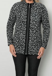 Donna che indossa una giacca grigia a maniche lunghe con stampa leopardata nera e una zip nera sul davanti, abbinata a pantaloni neri.