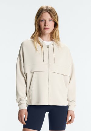 Vrouw met blond haar, gekleed in een beige hoodie met rits en marineblauwe fietsbroek, staand tegen een effen lichte achtergrond.