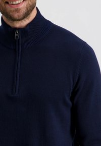 Navyfarbener Pullover mit hohem Rippkragen und halbem Reißverschluss. Glatte Strickstruktur mit sauberen Nähten und figurbetontem Schnitt.