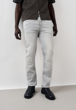 Muž v svetlosivých roztrhaných džínsach, čiernych kožených mokasínach a tmavej košeli so zipsom stojí pred jednoduchým svetlým pozadím.