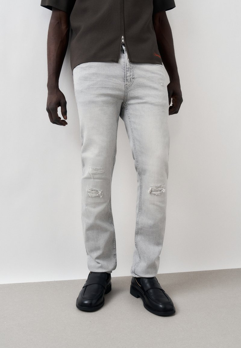 Άνδρας που φοράει ανοιχτό γκρι σκισμένα τζιν, μαύρα δερμάτινα μοκασίνια και σκούρο πουκάμισο με φερμουάρ, στέκεται μπροστά σε απλό φωτεινό φόντο.