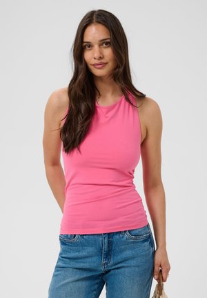 Femme aux longs cheveux foncés portant un top sans manches rose et un jean bleu, tenant un petit sac à main marron sur un fond uni.