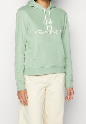 Personne vêtue d'un sweat à capuche GANT vert clair avec des cordons blancs et un pantalon couleur crème, debout devant un fond blanc uni.