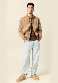 Chaqueta bomber marrón sobre una camisa marrón, vaqueros azul claro y zapatos de ante beige. Se lleva un bolso de hombro beige cruzado sobre el cuerpo.