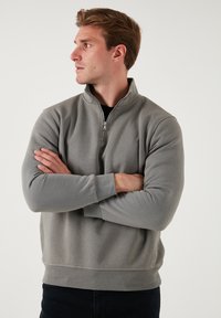 Felpa grigia con zip e colletto alto, dotata di maniche lunghe e una texture morbida, indossata sopra abbigliamento nero. Design minimalista senza motivi.