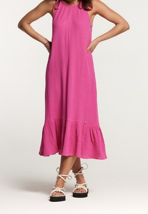 Femme en robe midi rose vif sans manches avec ourlet à volants, portant des sandales à plateforme blanches avec brides à la cheville, debout les mains sur les hanches.